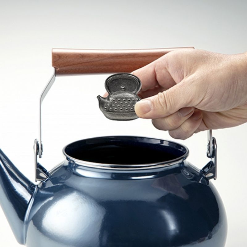 Hand holding Takaki Metal Cast Iron Egg for Nukadoko over a blue teapot with a wooden handle.