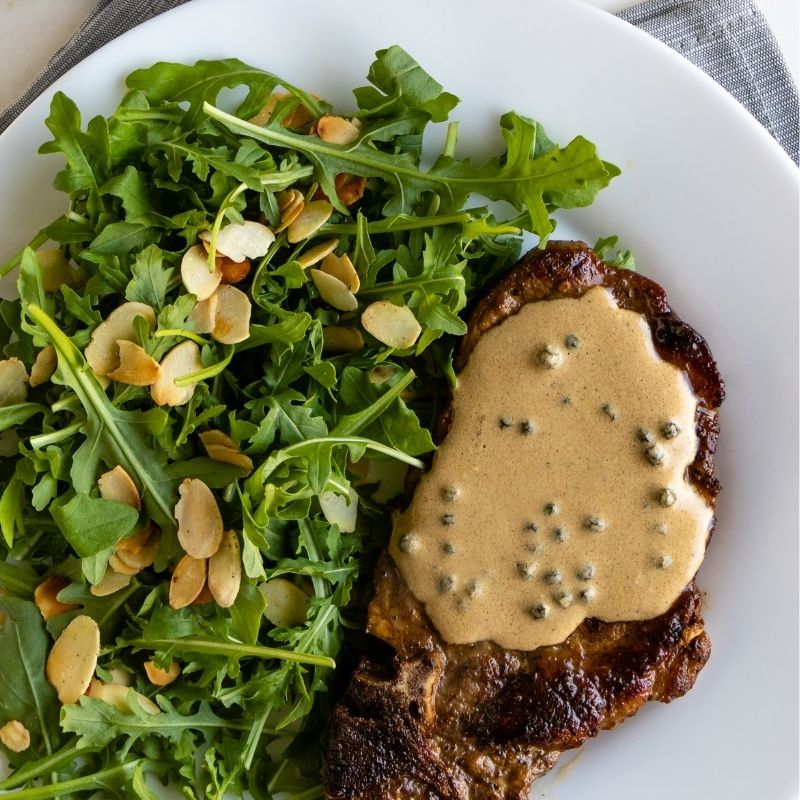 Steak with creamy Fresh Green Peppercorns sauce and arugula salad on a white plate
