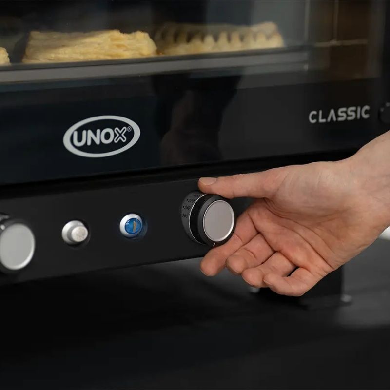 Close-up of a hand turning a control knob on a black Unox BAKERLUX CLASSIC MANUAL ELECTRIC Convection Oven - XENA-04HS-MADN, with baked pastries visible inside through a glass door.