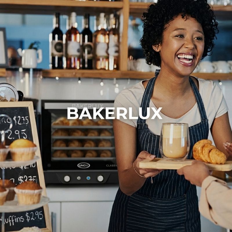 A smiling woman in a bakery wearing a dark navy pinstriped apron over a white t-shirt, holding a glass of coffee and croissants. The brand name 'BAKERLUX' is displayed in white text across the center of the image with Unox BAKERLUX CLASSIC MANUAL ELECTRIC Convection Oven - XENA-04HS-MADN in the background.