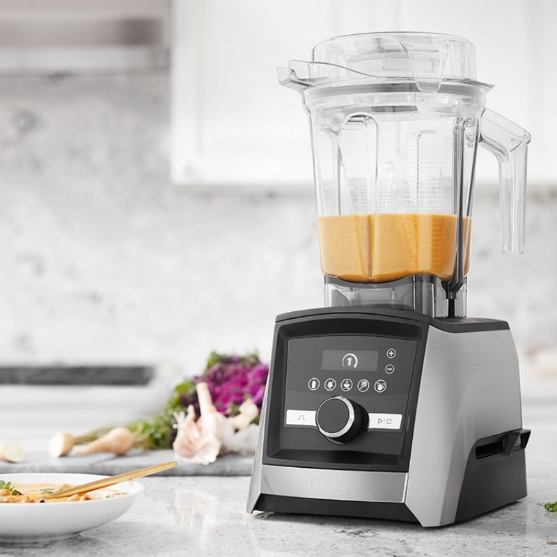 A Vitamix Ascent Series A3500i blender with a clear pitcher containing an orange-yellow liquid, set on a light stone kitchen counter with blurred vegetables and a bowl in the background.
