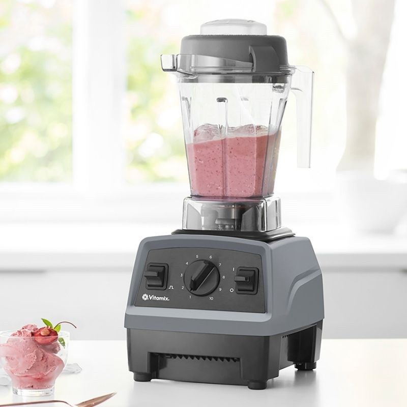 A Vitamix Explorian E310 Classic Blender with a gray base and clear jar, filled with a pink smoothie, set on a white countertop in front of a bright, softly blurred kitchen window. A small glass of pink dessert with a strawberry garnish sits to the left.