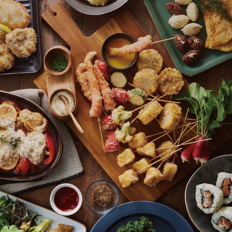 Assorted appetizers including fried skewers of kushi0katsu made with Wa Panko Gluten-Free Rice Panko Breadcrumbs, breaded items, and sushi on a wooden board.