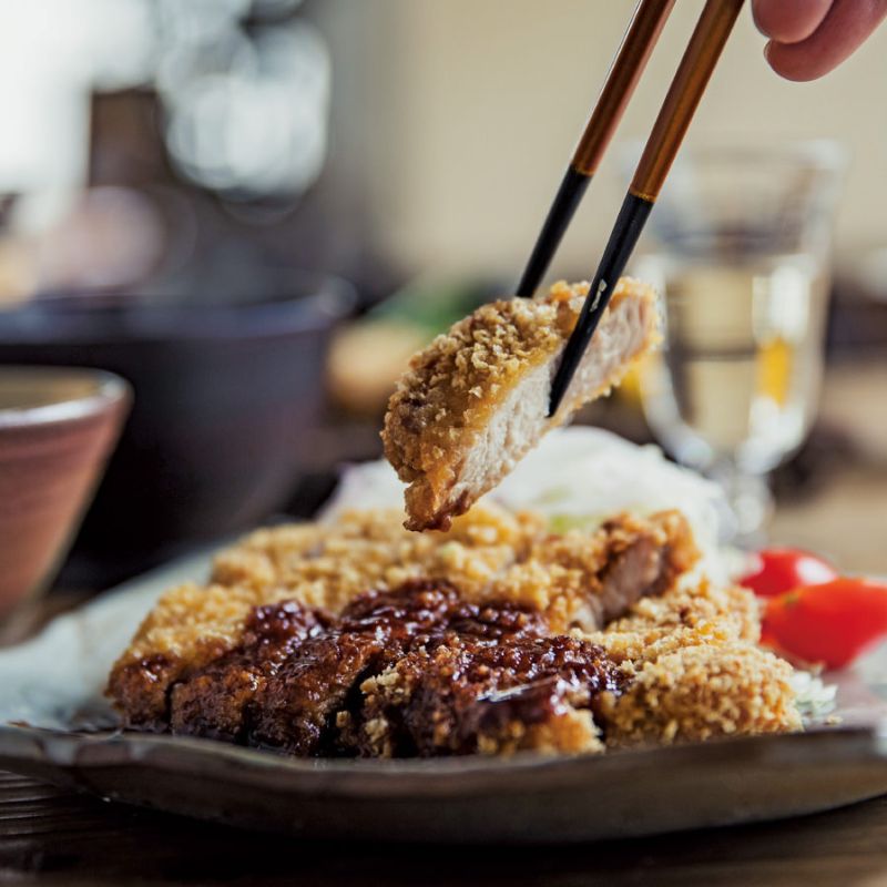 Fried pork cutlet made with Wa Panko Gluten-Free Rice Panko Breadcrumbs being held with chopsticks over a plate of similar cutlets on a blurred background.